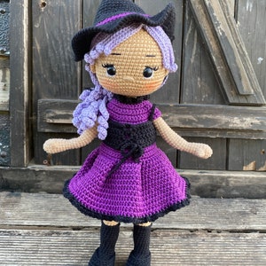 Pode incluir: Uma boneca de bruxa de croch&ecirc; com cabelo roxo, um chap&eacute;u preto e um vestido roxo. A boneca est&aacute; em p&eacute; sobre uma superf&iacute;cie de madeira.