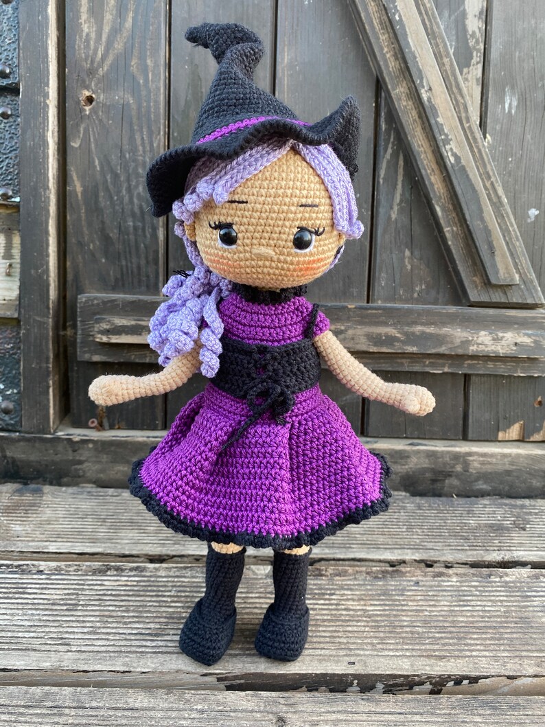 Pode incluir: Uma boneca de bruxa de croch&ecirc; com cabelo roxo, um chap&eacute;u preto e um vestido roxo. A boneca est&aacute; em p&eacute; sobre uma superf&iacute;cie de madeira.