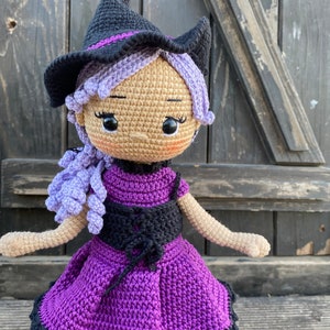 Pode incluir: Uma boneca de bruxa de croch&ecirc; com cabelo roxo, um chap&eacute;u preto e um vestido roxo com um corpete preto.
