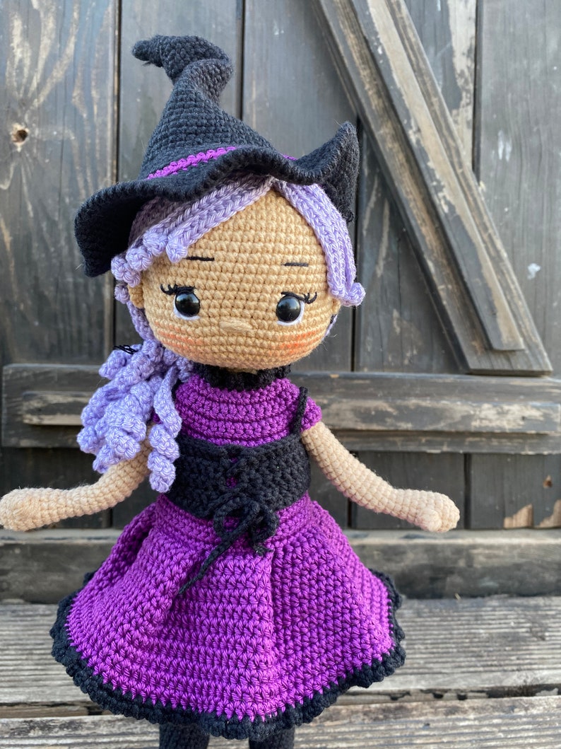 Pode incluir: Uma boneca de bruxa de croch&ecirc; com cabelo roxo, um chap&eacute;u preto e um vestido roxo com um corpete preto.