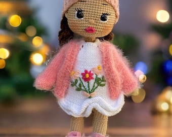 Puede incluir: Una muñeca de crochet con un gorro rosa con lazo, una chaqueta rosa, un vestido blanco con flores bordadas y zapatillas rosas y blancas.