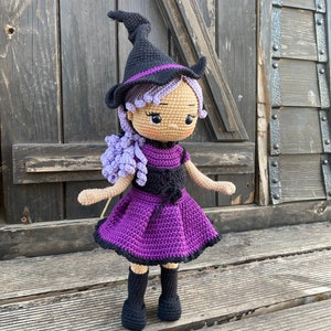 Pode incluir: Uma boneca de bruxa de croch&ecirc; com cabelo roxo, um chap&eacute;u preto e um vestido roxo. A boneca est&aacute; em p&eacute; sobre uma superf&iacute;cie de madeira.