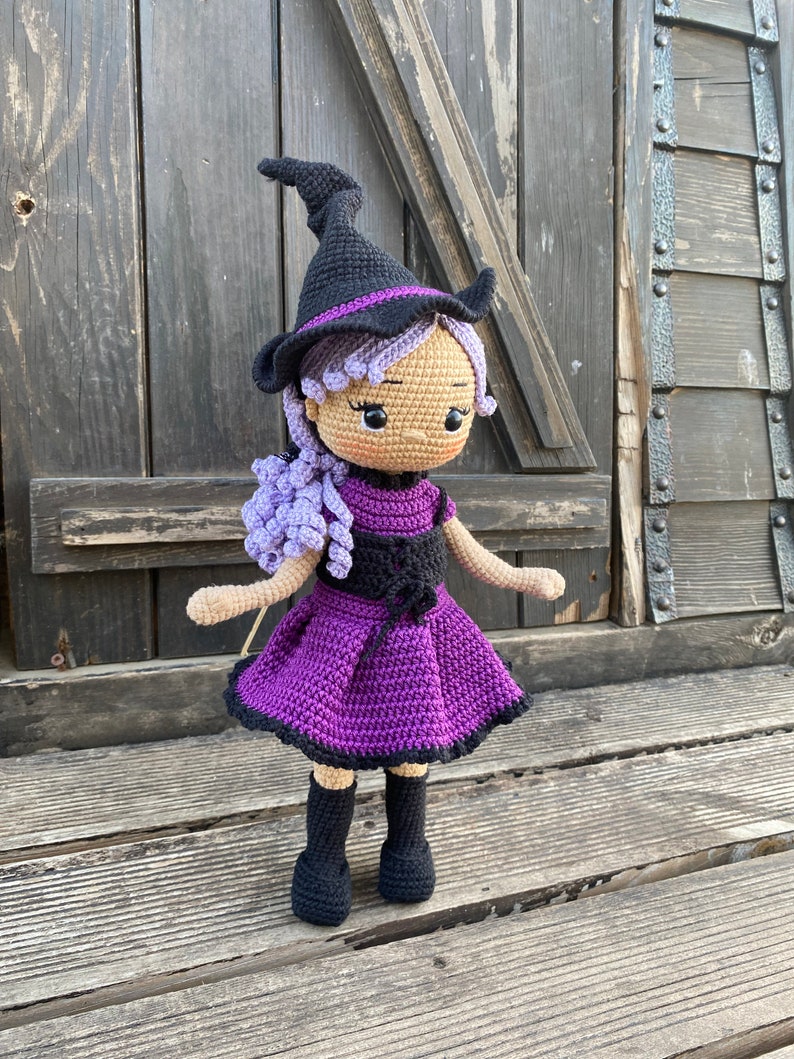 Pode incluir: Uma boneca de bruxa de croch&ecirc; com cabelo roxo, um chap&eacute;u preto e um vestido roxo. A boneca est&aacute; em p&eacute; sobre uma superf&iacute;cie de madeira.