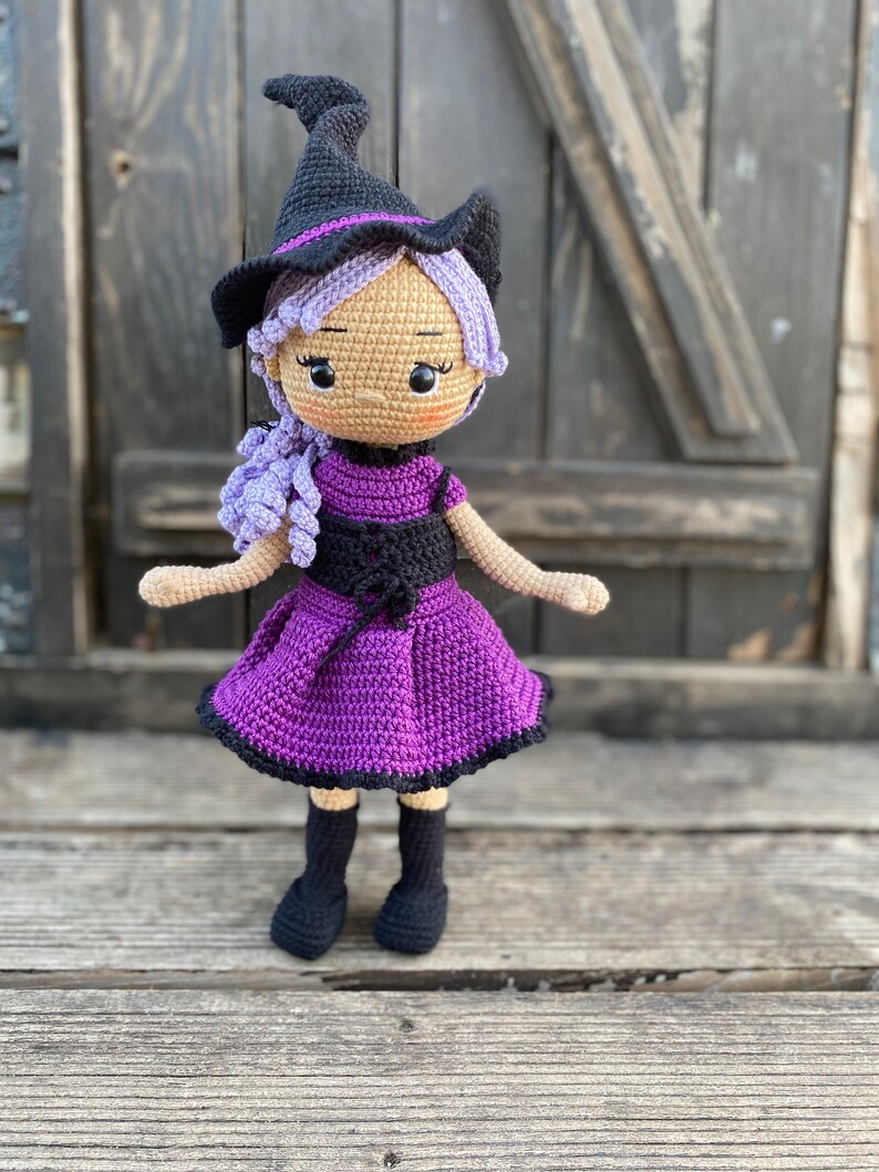 Pode incluir: Uma boneca de bruxa de croch&ecirc; com cabelo roxo, um chap&eacute;u preto e um vestido roxo. A boneca est&aacute; em p&eacute; sobre uma superf&iacute;cie de madeira.