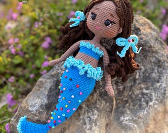 Puede incluir: Una muñeca de sirena de crochet con cabello castaño, ojos azules y una cola azul con cuentas de colores. La muñeca lleva un top azul y está sentada en una roca.