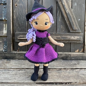 Pode incluir: Uma boneca de bruxa de croch&ecirc; com cabelo roxo, um chap&eacute;u preto e um vestido roxo. A boneca est&aacute; em p&eacute; sobre uma superf&iacute;cie de madeira.