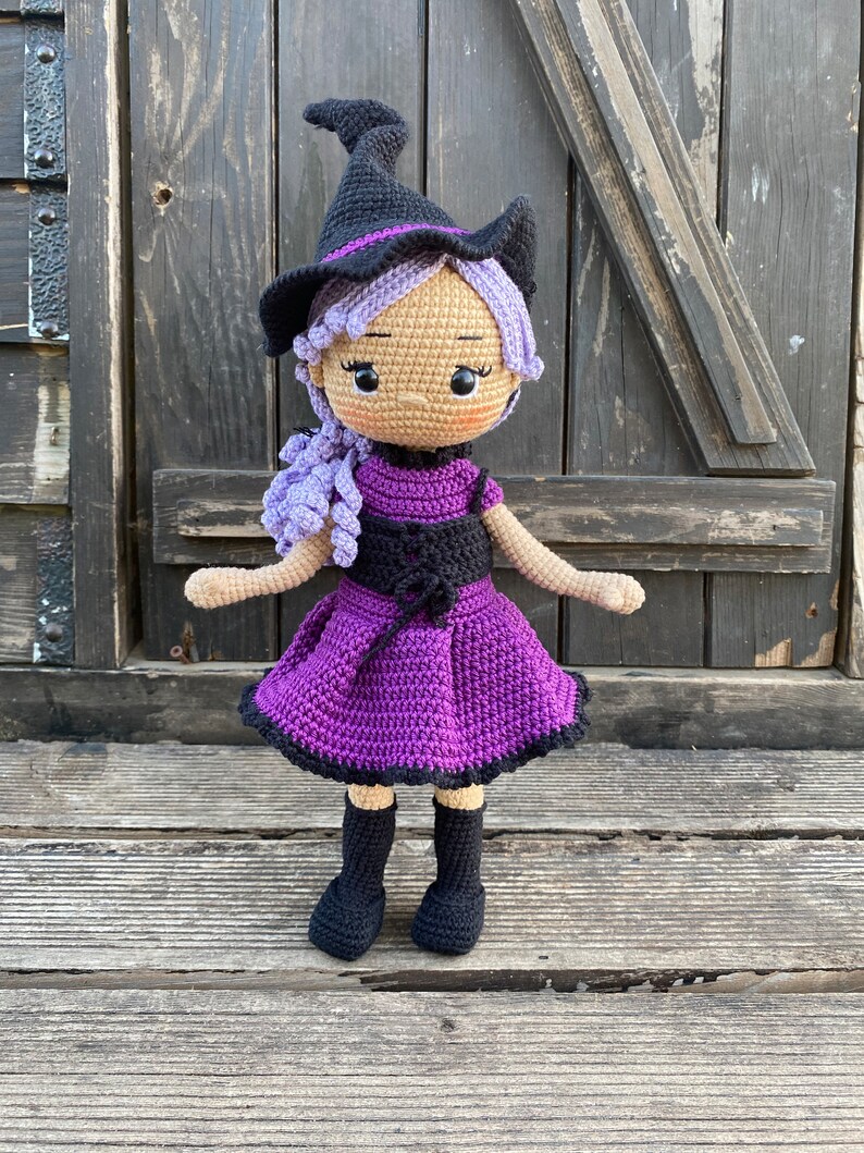 Pode incluir: Uma boneca de bruxa de croch&ecirc; com cabelo roxo, um chap&eacute;u preto e um vestido roxo. A boneca est&aacute; em p&eacute; sobre uma superf&iacute;cie de madeira.