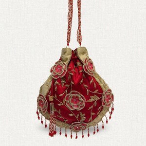Peut inclure: Un sac rouge en satin avec cordon de serrage, garniture dorée et broderies florales. Le sac est orné de poignées perlées et de perles rouges pendantes. Les fleurs sont rouges avec des accents dorés.