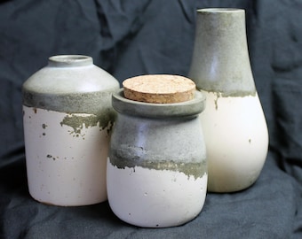 May include: Three ceramic vases in a neutral color palette. The vases are white with a gray-green band and a distressed finish. One vase has a cork stopper. The shapes vary, including a jar, a bottle, and a vase.