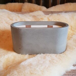 May include: A grey concrete candle holder with a white wax interior and two wooden wicks. The oval container sits on a fluffy, cream-coloured surface. A modern home decor item.