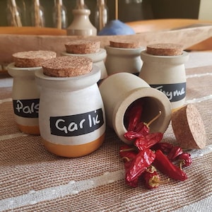 Può includere: Sei piccoli vasetti di ceramica bianca con tappi di sughero ed etichette nere. Le etichette sono scritte a mano con le parole "Prezzemolo", "Aglio", "Timo". Un vasetto è aperto e pieno di peperoncini rossi.