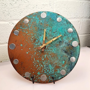 May include: A round, copper clock with a turquoise patina and gold hands. The clock face has silver metal accents around the edge.