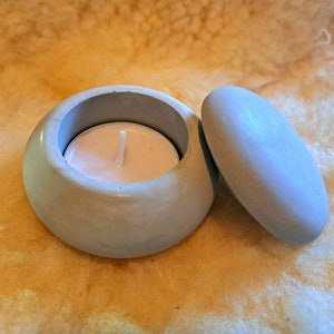 May include: A light grey concrete candle holder with a lid. The holder has a small white tea light candle inside.