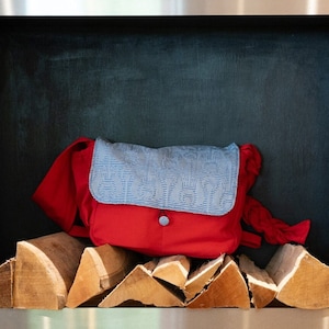 May include: A red and blue fabric bag with a flap closure sits on a pile of wood logs in a metal frame.