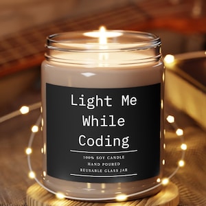Może przedstawiać: Brązowa świeca sojowa z czarną etykietą, na której widnieje napis "Light Me While Coding". Etykieta zawiera również informacje: "100% Soy Candle, Hand Poured, Reusable Glass Jar". Świeca stoi na drewnianym podkładce i jest otoczona białymi światełkami.