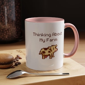 May include: White ceramic mug with a pink rim and handle. The mug has a pixelated image of a brown and white cow and the text "Thinking About My Farm".