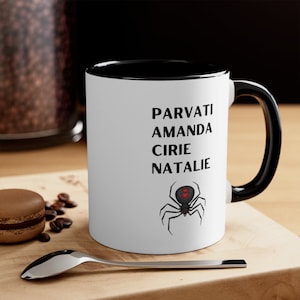 May include: White ceramic mug with a black rim and handle. The mug has black text that reads "Parvati Amanda Cirie Natalie" and a black spider with red eyes.