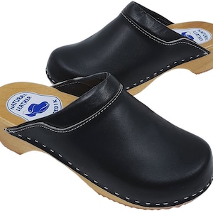 Peut inclure: Paire de sabots en cuir noir avec semelles en bois et une bordure blanche cousue. Les sabots portent une étiquette indiquant "Cuir Naturel".