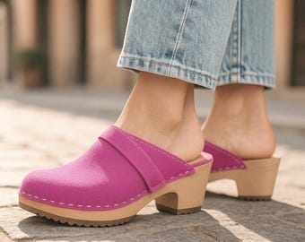 May include: Bright pink clogs with a wooden heel and sole. The clogs have a strap across the top and are accented with white stitching. The shoes are worn with blue jeans.