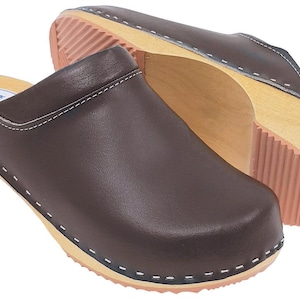 Può includere: Paio di zoccoli in pelle marrone con suole in legno e battistrada in gomma rosa. Gli zoccoli hanno la punta arrotondata e sono cuciti con filo bianco. Le parole "NATURAL LEATHER" sono stampate sulla soletta.