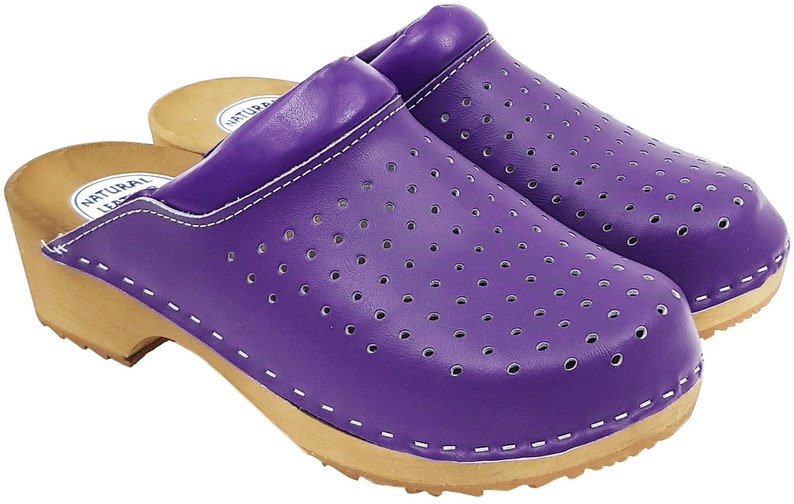 May include: Purple leather clogs with a wooden sole and a perforated design. The clogs have a white stitching detail around the edge of the sole and a label that says "NATURAL LEATHER" on the side.