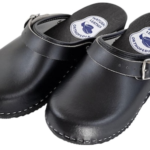 Può includere: Un paio di zoccoli in pelle nera con cinturino con fibbia. Gli zoccoli hanno cuciture bianche attorno alla suola e una soletta in pelle naturale con il testo "NATURAL LEATHER" e "ORTHOPEDIK".