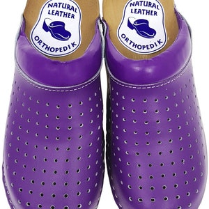 May include: Pair of purple leather clogs with a perforated design and a wooden base. The clogs have a label that reads "Natural Leather OrthopediK".