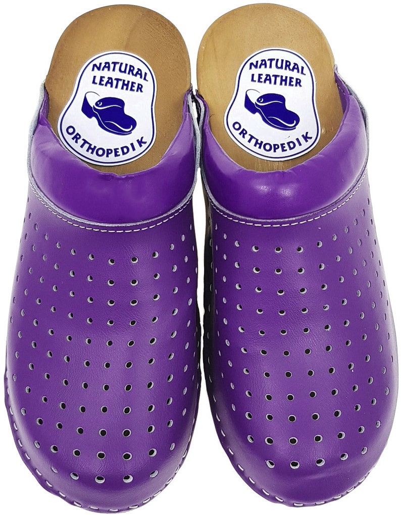 May include: Pair of purple leather clogs with a perforated design and a wooden base. The clogs have a label that reads "Natural Leather OrthopediK".