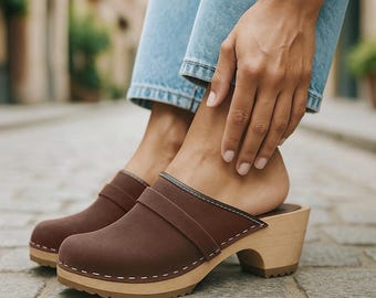 May include: Brown clogs with a wooden sole and a strap across the top. The clogs have a rounded toe and a heel. The sole is light brown with a dark brown rubber tread. The clogs are worn with blue jeans.