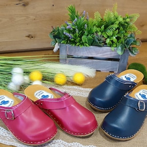 Könnte beinhalten: Vier Paar Ledersandalen mit Holzsohlen. Zwei Paar sind rosa und zwei Paar sind blau. Jedes Paar hat einen Schnallenriemen und ein Etikett mit der Aufschrift "Naturleder".