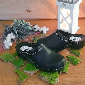Può includere: Un paio di zoccoli in pelle nera con un dettaglio di cucitura bianca. Gli zoccoli hanno una suola in legno e sono appoggiati su un letto di muschio verde.