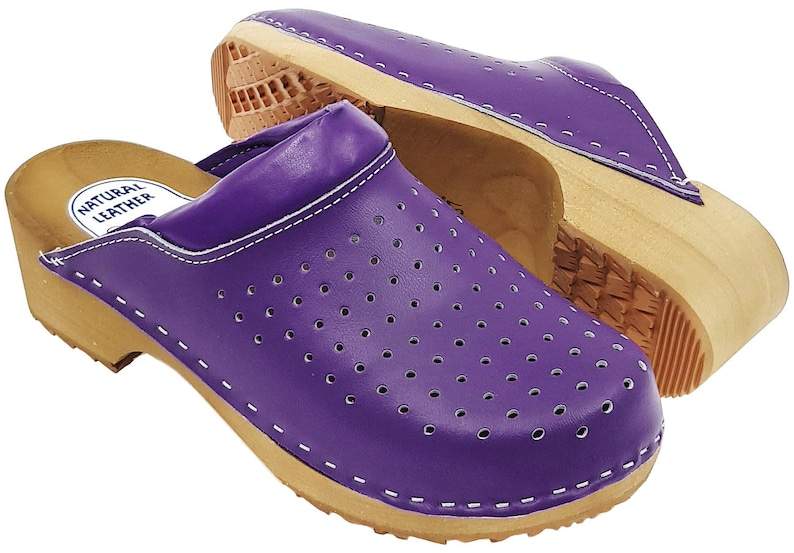 May include: Purple leather clogs with a wooden sole and a white stitch detail. The clogs have a perforated design and the words "Natural Leather" are printed on the inside of the clog.