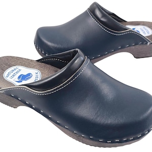 May include: A pair of navy blue leather clogs with wooden soles. The clogs have white stitching around the edges and a black trim. The insoles are stamped with "NATURAL LEATHER" and a logo. These clogs are a type of shoe.