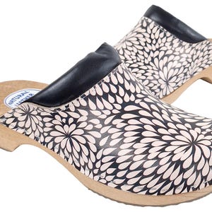 May include: A pair of clogs with a floral pattern. The clogs have a wooden sole and a black leather upper. The floral pattern is white on a black background. The clogs are designed for comfort and style.