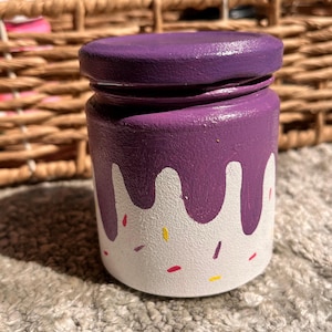 May include: A small, hand-painted jar with a purple lid and a white base. The jar has a purple dripping design and colourful sprinkles. The jar is on a grey, textured surface.