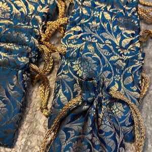 Könnte beinhalten: Eine Sammlung kleiner, verzierter Beutel mit Kordelzug. Die Beutel sind tief türkisblau mit einem goldenen Blumenmuster. Jeder Beutel hat einen goldenen Kordelverschluss.