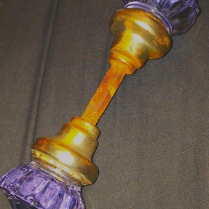 May include: A door knob set featuring two fluted, translucent purple glass knobs attached to a central, weathered, rust-colored metal spindle. Each glass knob is secured by a tarnished brass-toned metal base, creating a contrast between the vibrant purple glass and the aged, oxidized metal components of the hardware assembly.