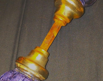May include: A door knob set featuring two fluted, translucent purple glass knobs attached to a central, weathered, rust-colored metal spindle. Each glass knob is secured by a tarnished brass-toned metal base, creating a contrast between the vibrant purple glass and the aged, oxidized metal components of the hardware assembly.