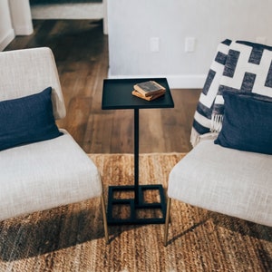 May include: Two white chairs with blue pillows and a black side table with a stack of books on top. The chairs and table are on a brown rug.