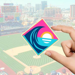Pode incluir: Um adesivo quadrado com um design estilizado de onda, sol e oceano em rosa, azul-petr&oacute;leo e branco. O adesivo &eacute; segurado em frente a um est&aacute;dio de beisebol com um horizonte da cidade ao fundo.