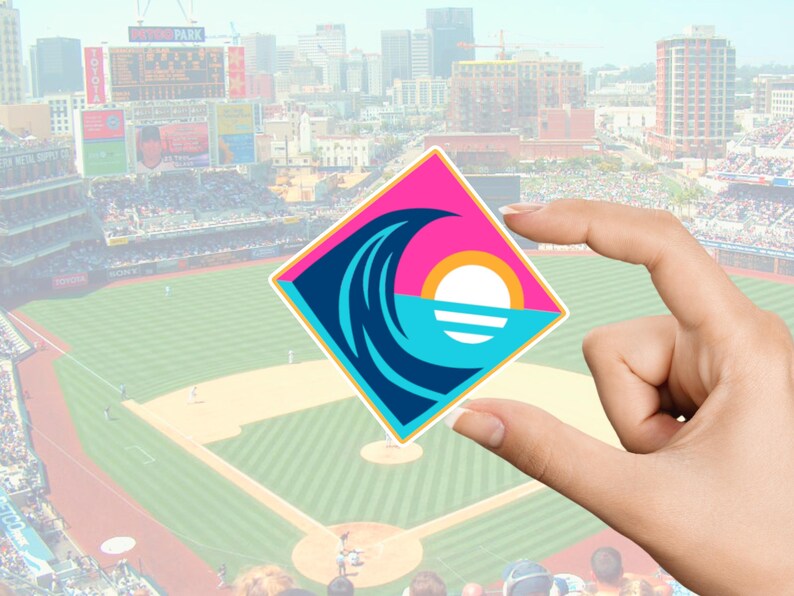 Pode incluir: Um adesivo quadrado com um design estilizado de onda, sol e oceano em rosa, azul-petr&oacute;leo e branco. O adesivo &eacute; segurado em frente a um est&aacute;dio de beisebol com um horizonte da cidade ao fundo.