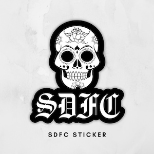 May include: Black and white sticker featuring a sugar skull design with floral accents. The sticker has the letters "SDFC" in a gothic font, with the words "SDFC STICKER" below.