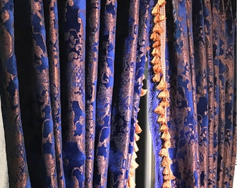 May include: Floor-length curtains featuring a deep blue fabric with a raised copper-coloured damask pattern. The vertical edges of the curtains are finished with a decorative trim consisting of a light brown woven band and small hanging tassels.