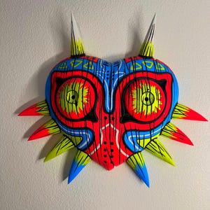 May include: A colorful, heart-shaped mask with a striking design. The mask features a vibrant palette of red, blue, yellow, and black. It has two large, circular eye designs and multiple pointed protrusions. The mask is mounted on a white wall.