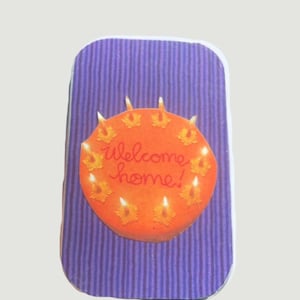 May include: A rectangular card with a purple striped background. An orange circle with the words "welcome home!" is centered on the card. The circle has yellow flower-shaped candles around its perimeter.