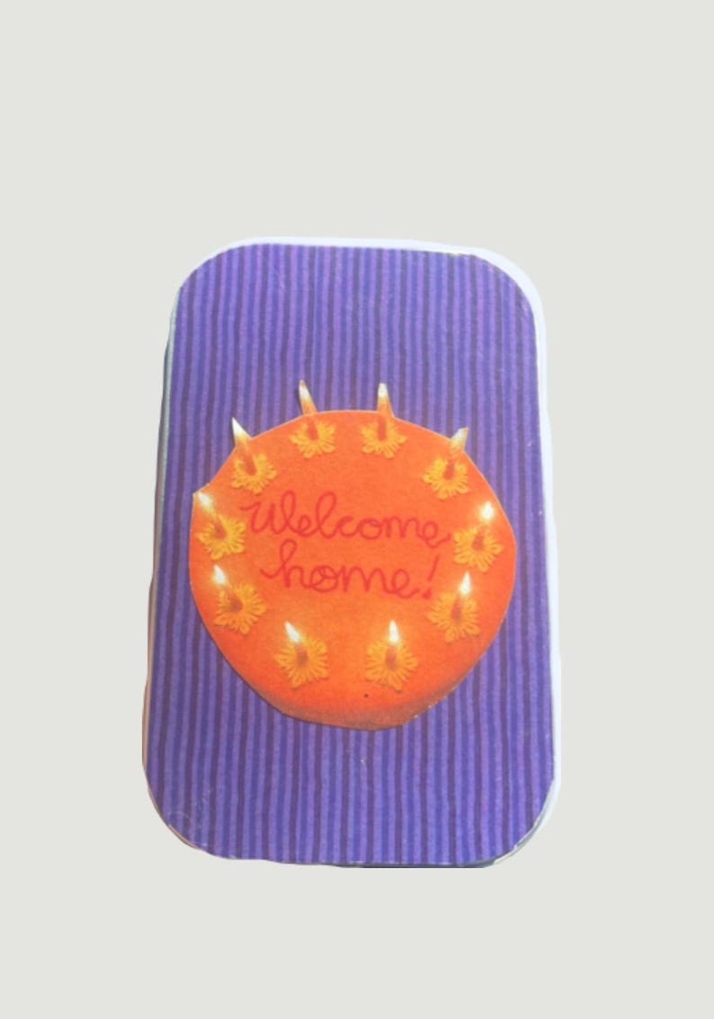 May include: A rectangular card with a purple striped background. An orange circle with the words "welcome home!" is centered on the card. The circle has yellow flower-shaped candles around its perimeter.