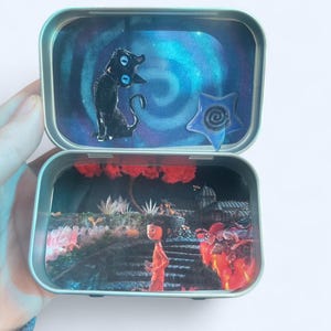 May include: Open metal tin with two compartments, each featuring a different scene. The top compartment has a black cat figure and a blue star against a swirling blue background. The bottom compartment shows a person in an orange outfit in a garden.