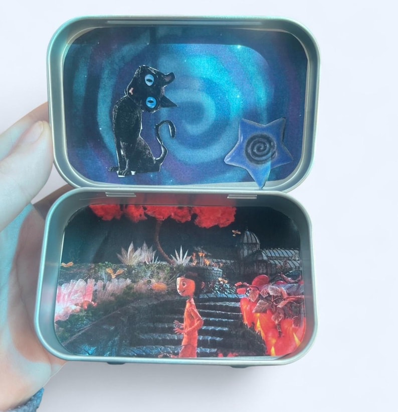 May include: Open metal tin with two compartments, each featuring a different scene. The top compartment has a black cat figure and a blue star against a swirling blue background. The bottom compartment shows a person in an orange outfit in a garden.
