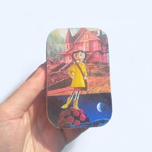 May include: A rectangular tin featuring a cartoon illustration of a girl in a yellow coat and boots, standing in front of a pink house. The background includes a night sky with a moon and a daytime scene with a house.