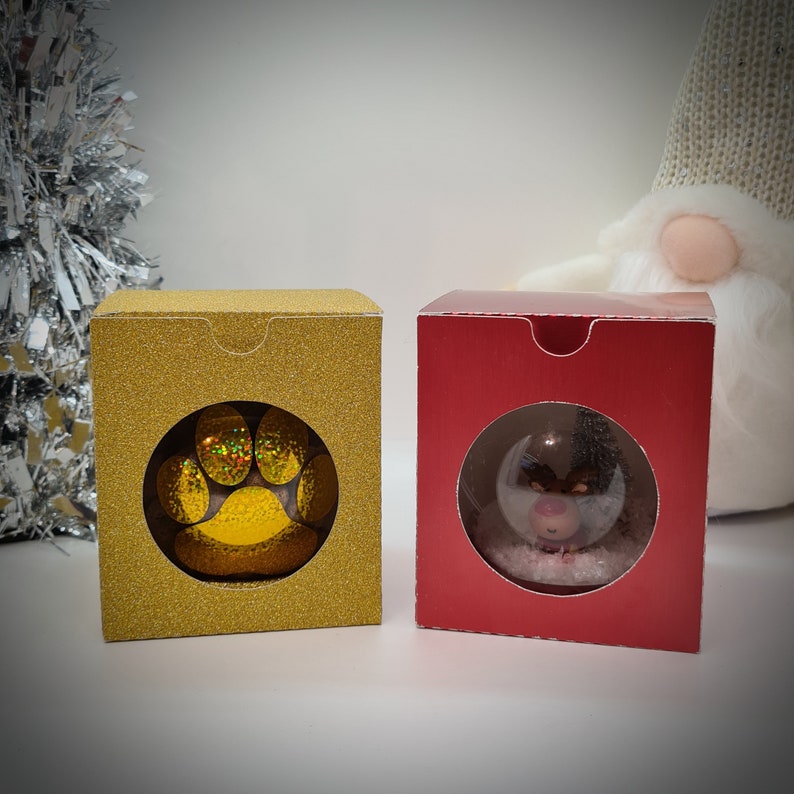 Op de afbeelding: Twee decoratieve dozen met een venster om een klein ornament te tonen. De doos aan de linkerkant is goud met een glitterafwerking en heeft een holografische pootafdruk. De doos aan de rechterkant is rood met een glitterafwerking en heeft een sneeuwbol met een klein dier erin.